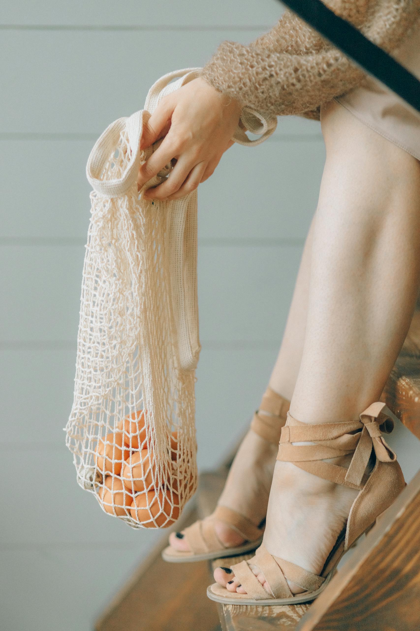 Elegant woman sitting on stairs holding a mesh bag containing oranges, showcasing sustainable fashion.