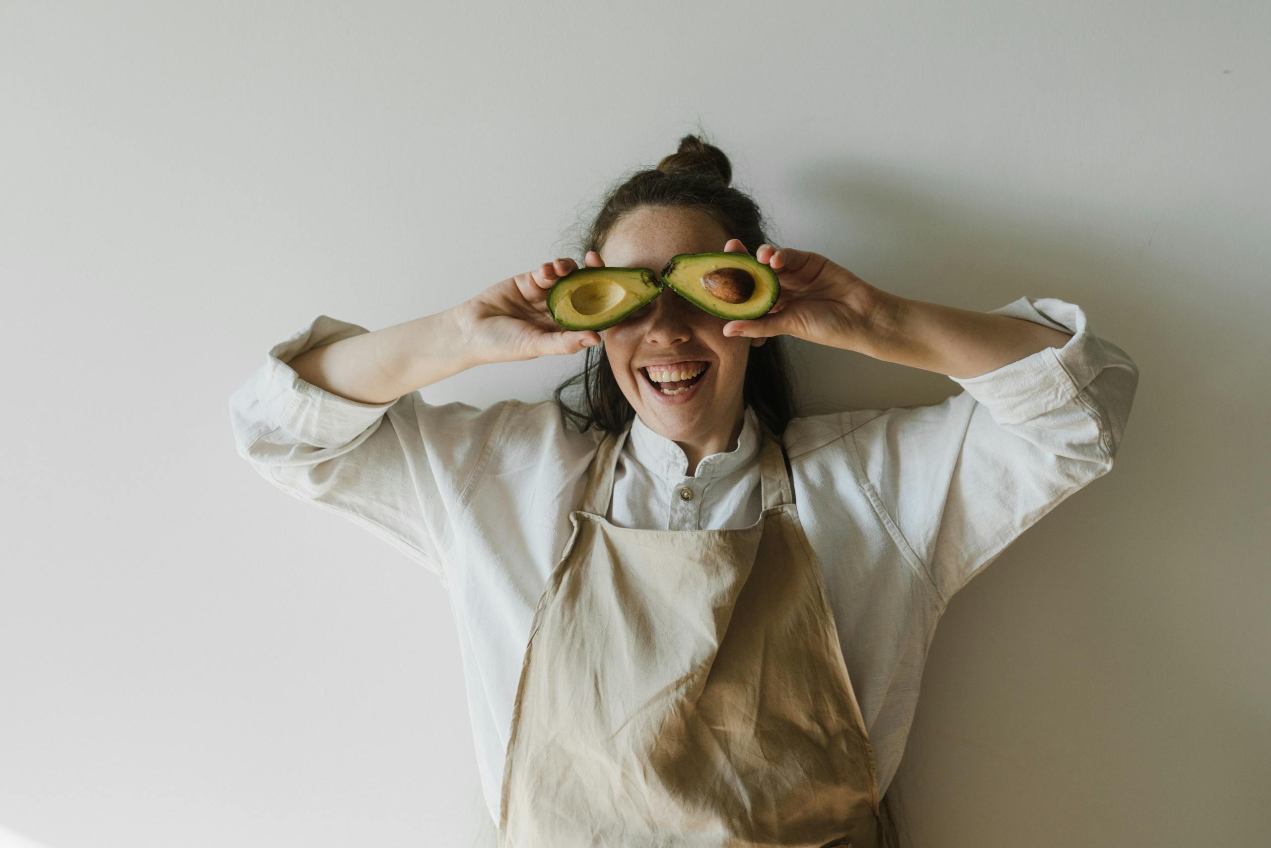 Joyful woman holding avocado halves over her eyes, symbolizing healthy living and fun creativity.