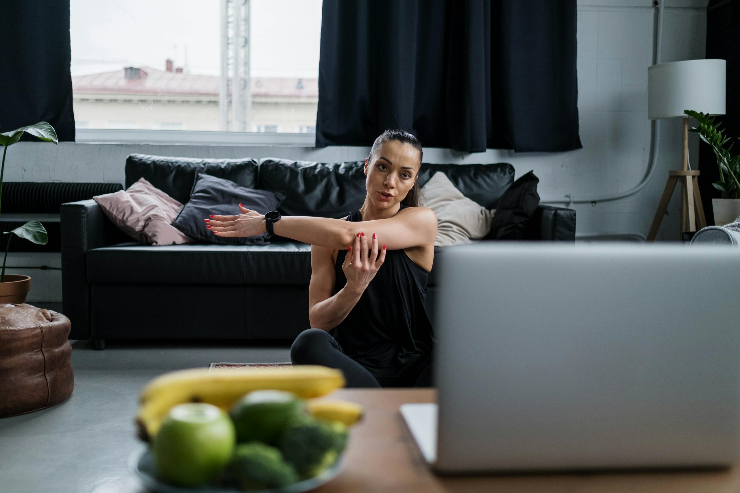 Woman practicing yoga at home with a laptop, stretching arms in a cozy living room setting.