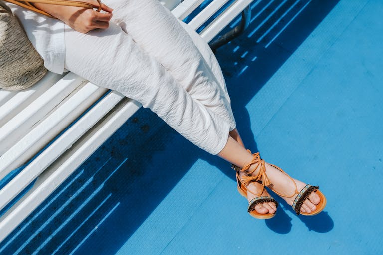 A woman sitting on a bench outdoors, showcasing stylish sandals and white pants on a sunny day.