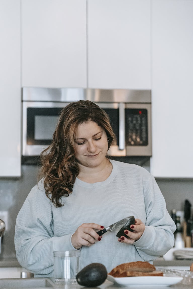 Optimistic female cook cutting ripe avocado while making toasts in modern light kitchen with microwave oven at home during breakfast