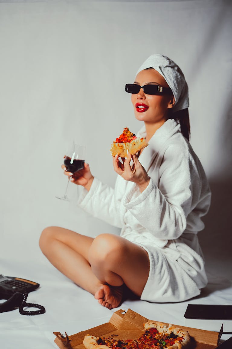 Stylish woman in robe enjoying pizza and wine, embracing a luxury lifestyle.