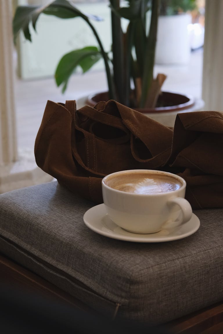 Warm cappuccino on a seat with a brown tote bag beside it in a cozy cafe setting.