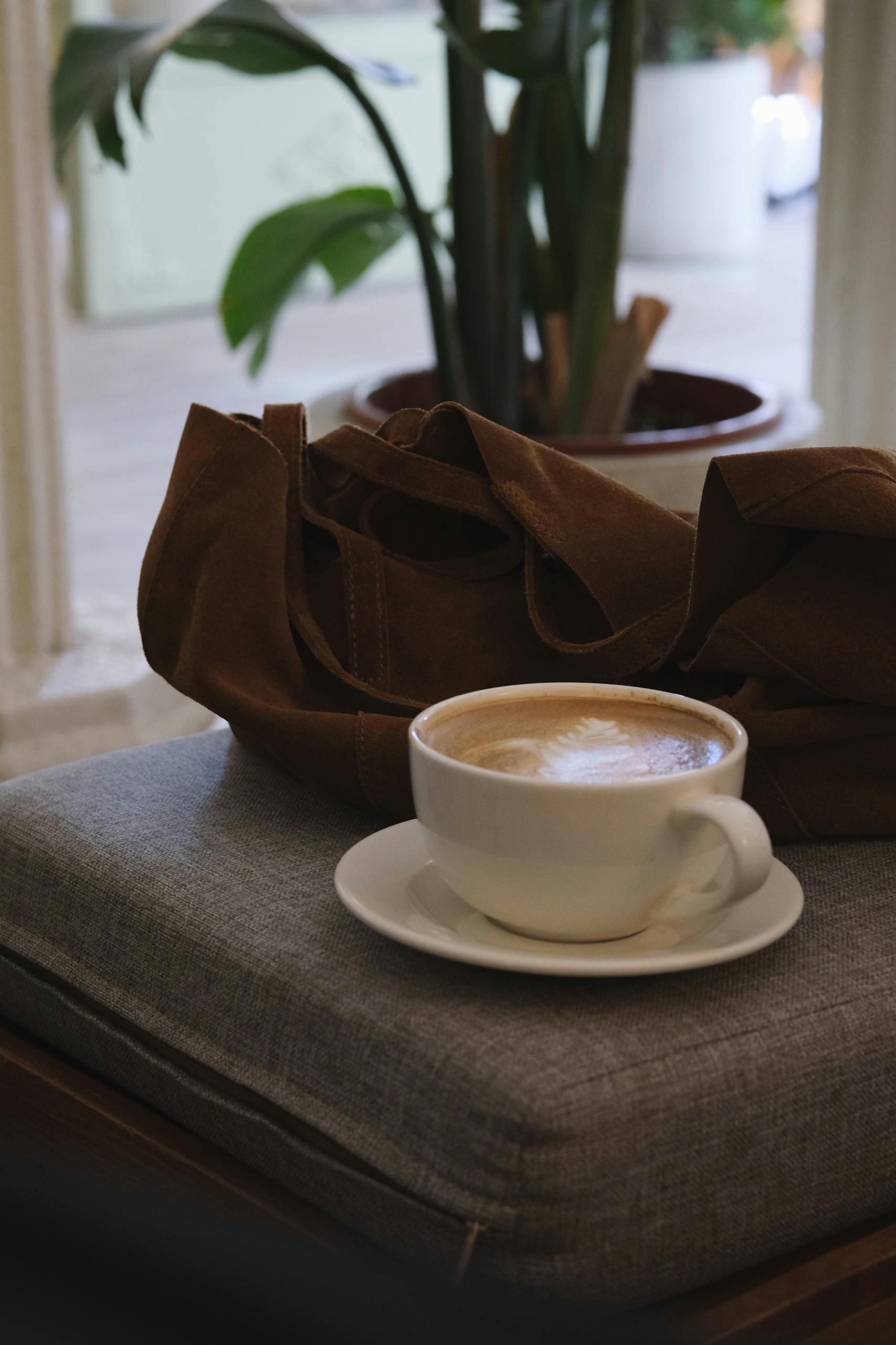 Warm cappuccino on a seat with a brown tote bag beside it in a cozy cafe setting.