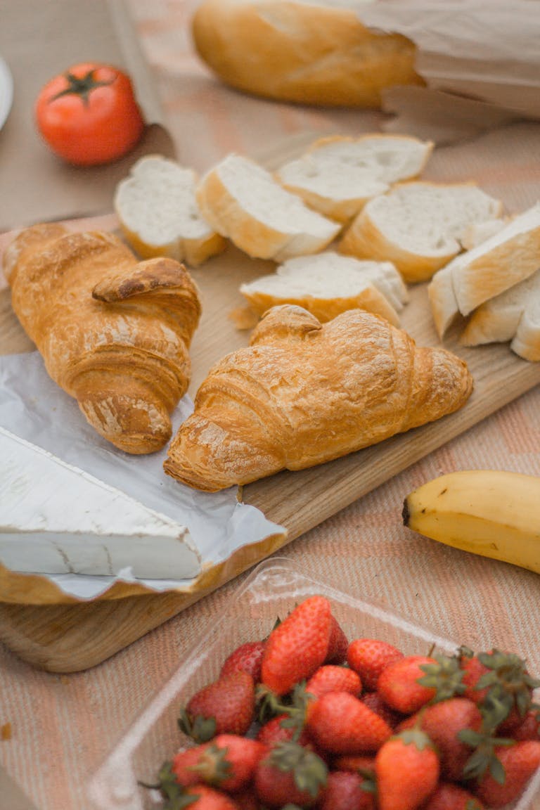 A picnic setup featuring croissants, strawberries, banana, tomato, bread slices, and Brie cheese.