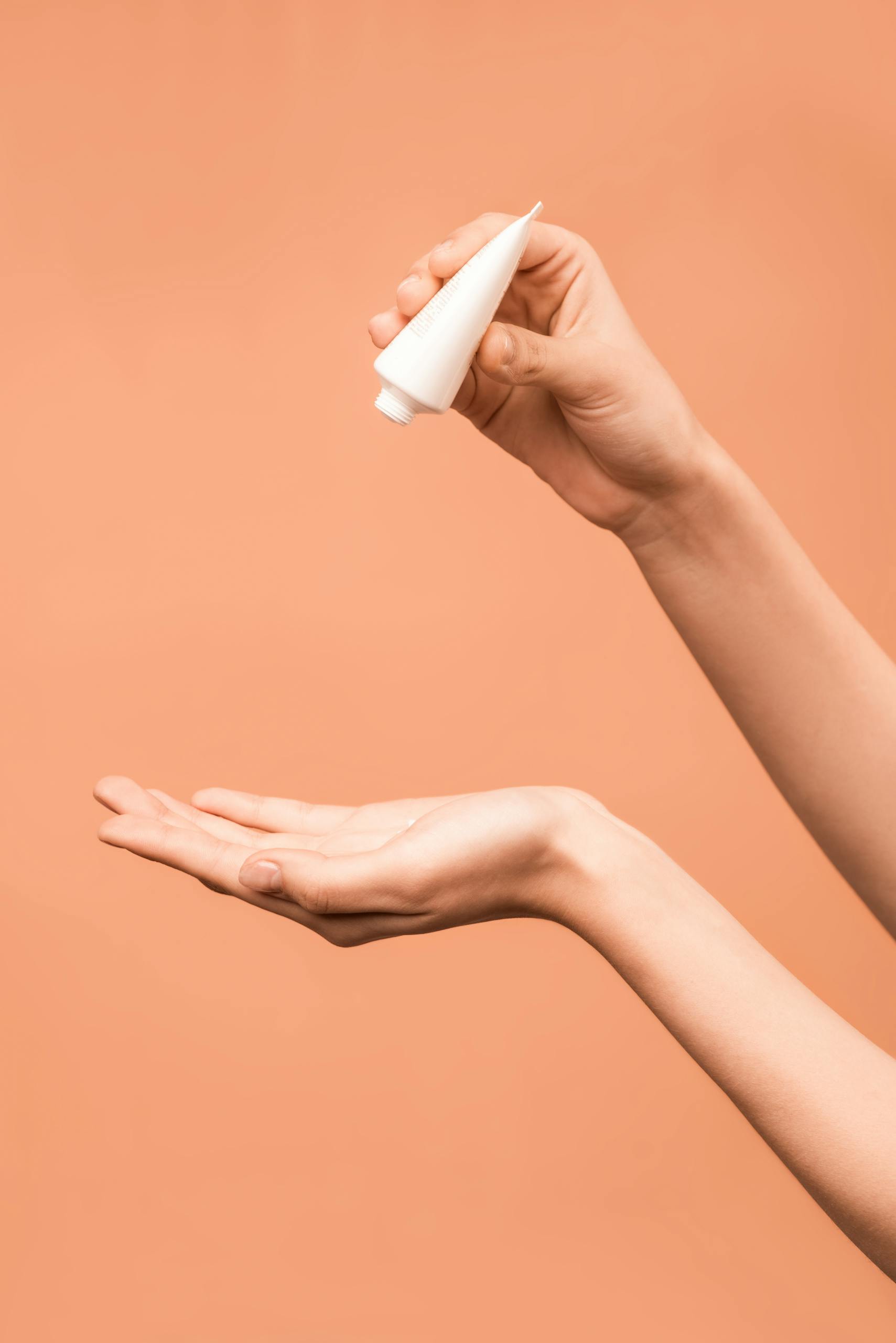 Close-up of hands applying lotion from a tube against a peach background.