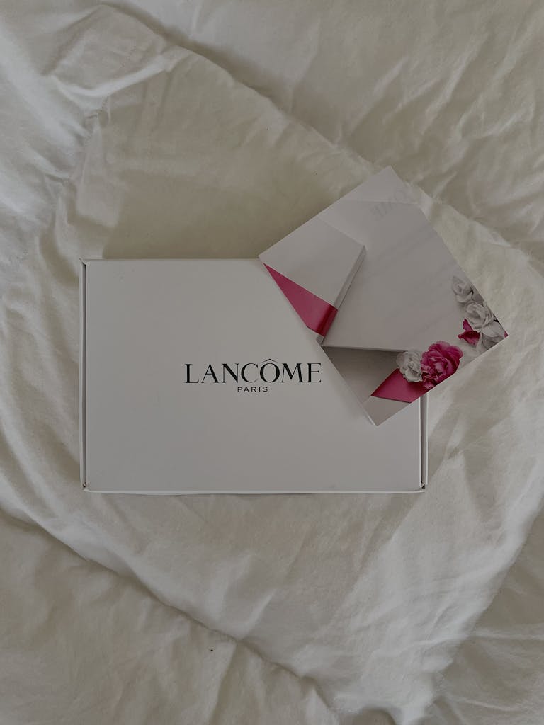Top view of Lancome gift box and floral envelope on a white bed.