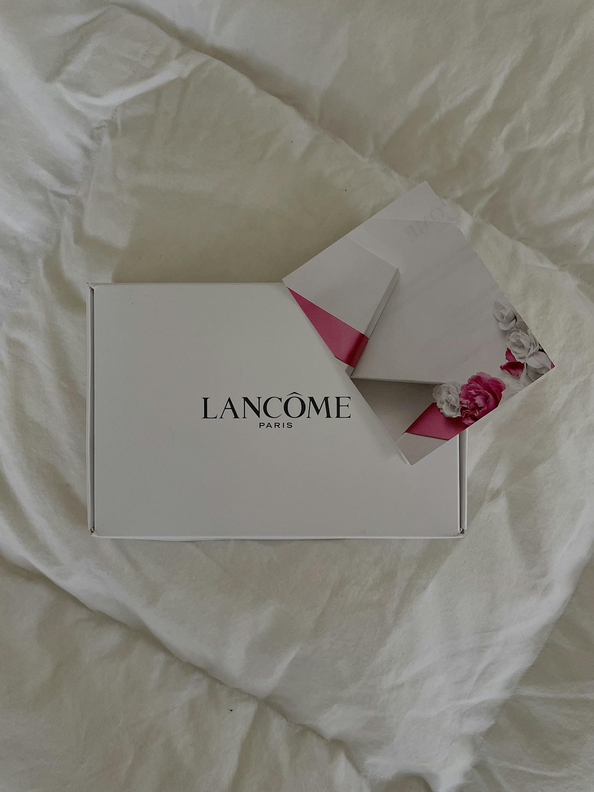 Top view of Lancome gift box and floral envelope on a white bed.