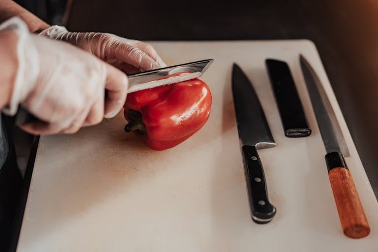 A person wearing gloves slices a red bell pepper on a cutting board using sharp knives.