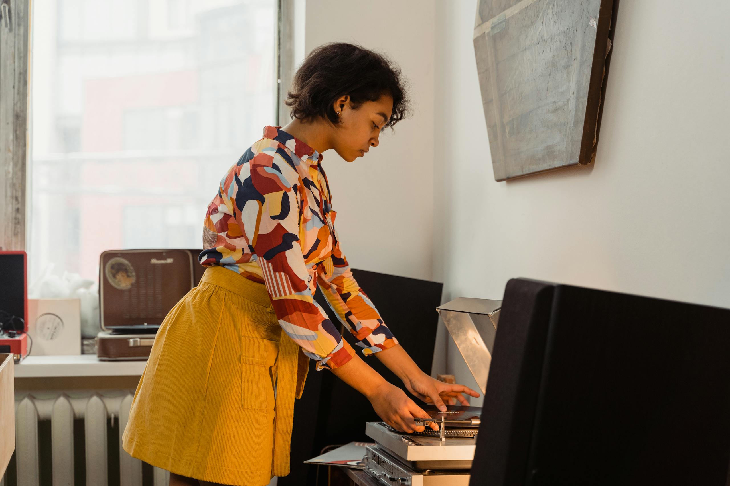 A woman in a colorful printed shirt and yellow skirt using a vintage vinyl record player indoors.
