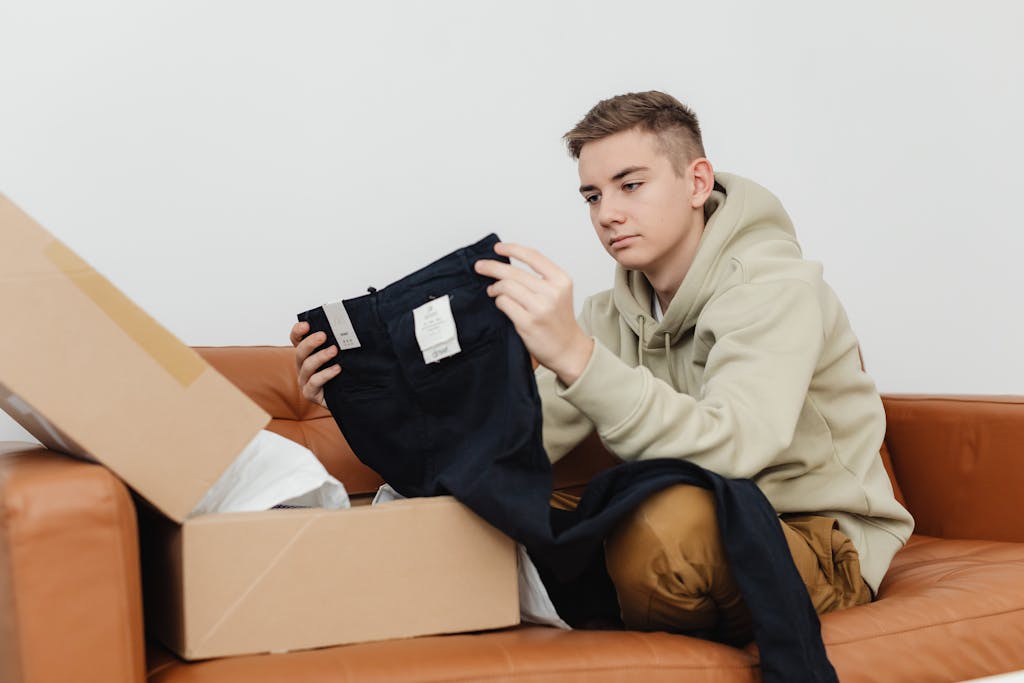 Teen sitting on sofa unboxing new clothes from a cardboard box.