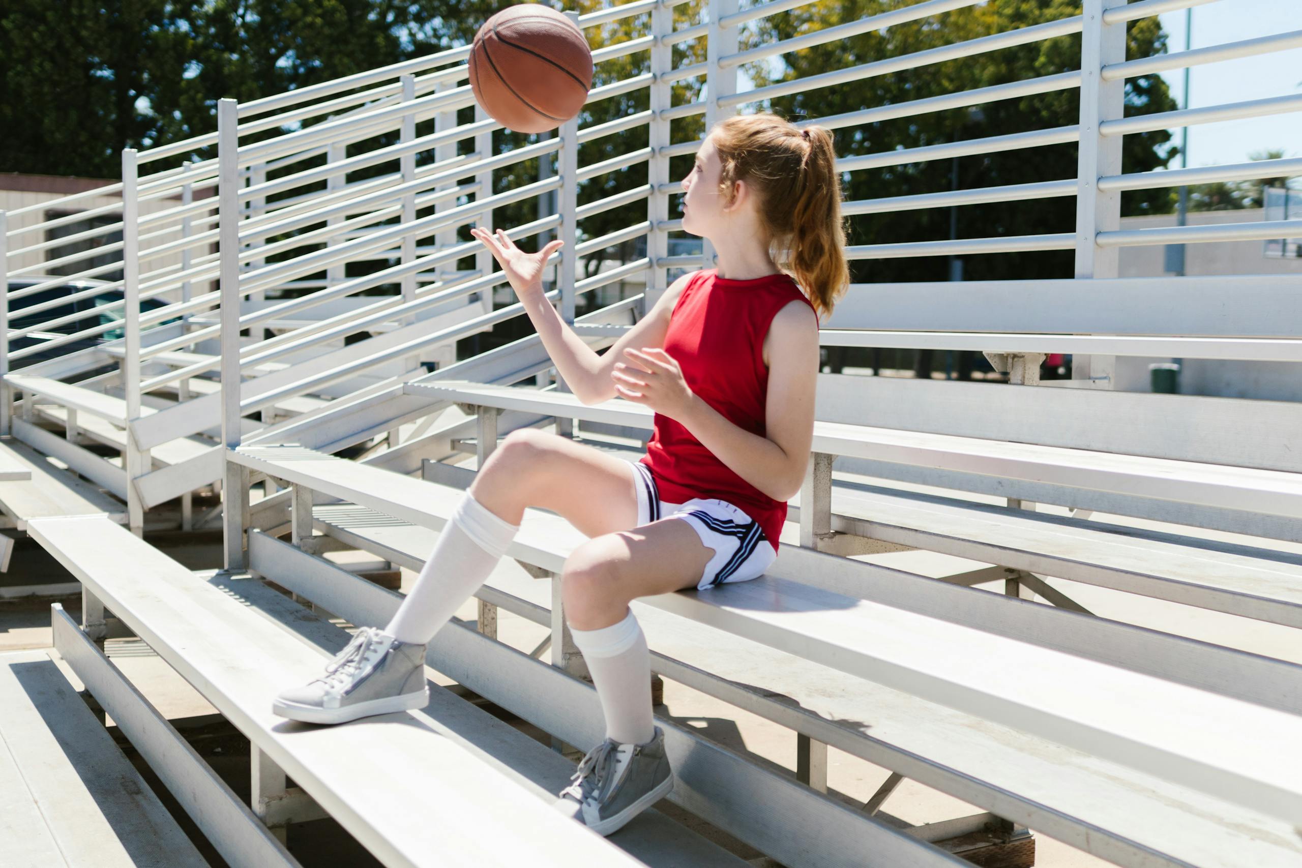 A young girl in sportswear sitting on bleachers outdoors, playing with a basketball.
