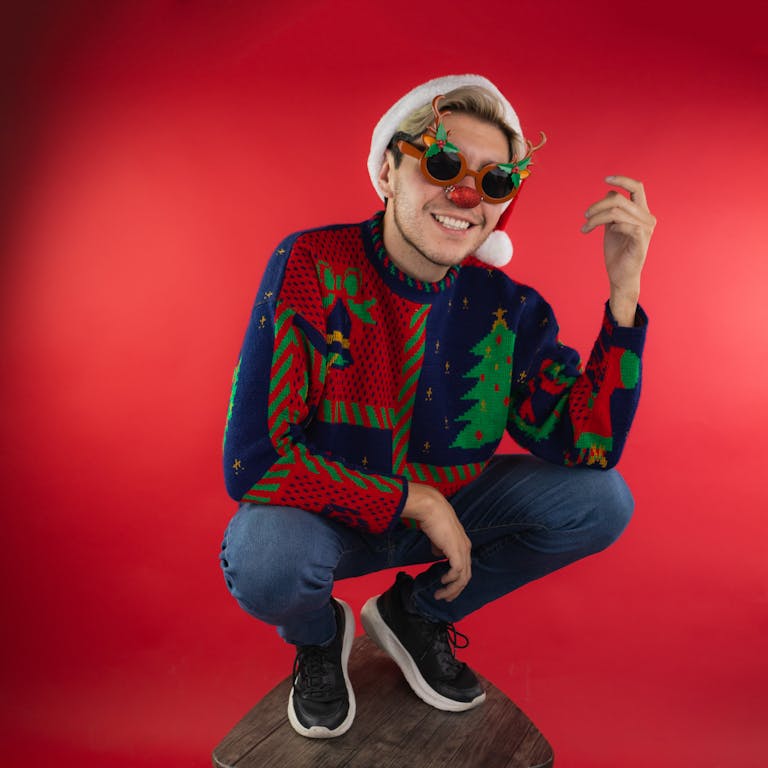 Playful young adult wearing a Christmas sweater and festive glasses, posing against a red background.