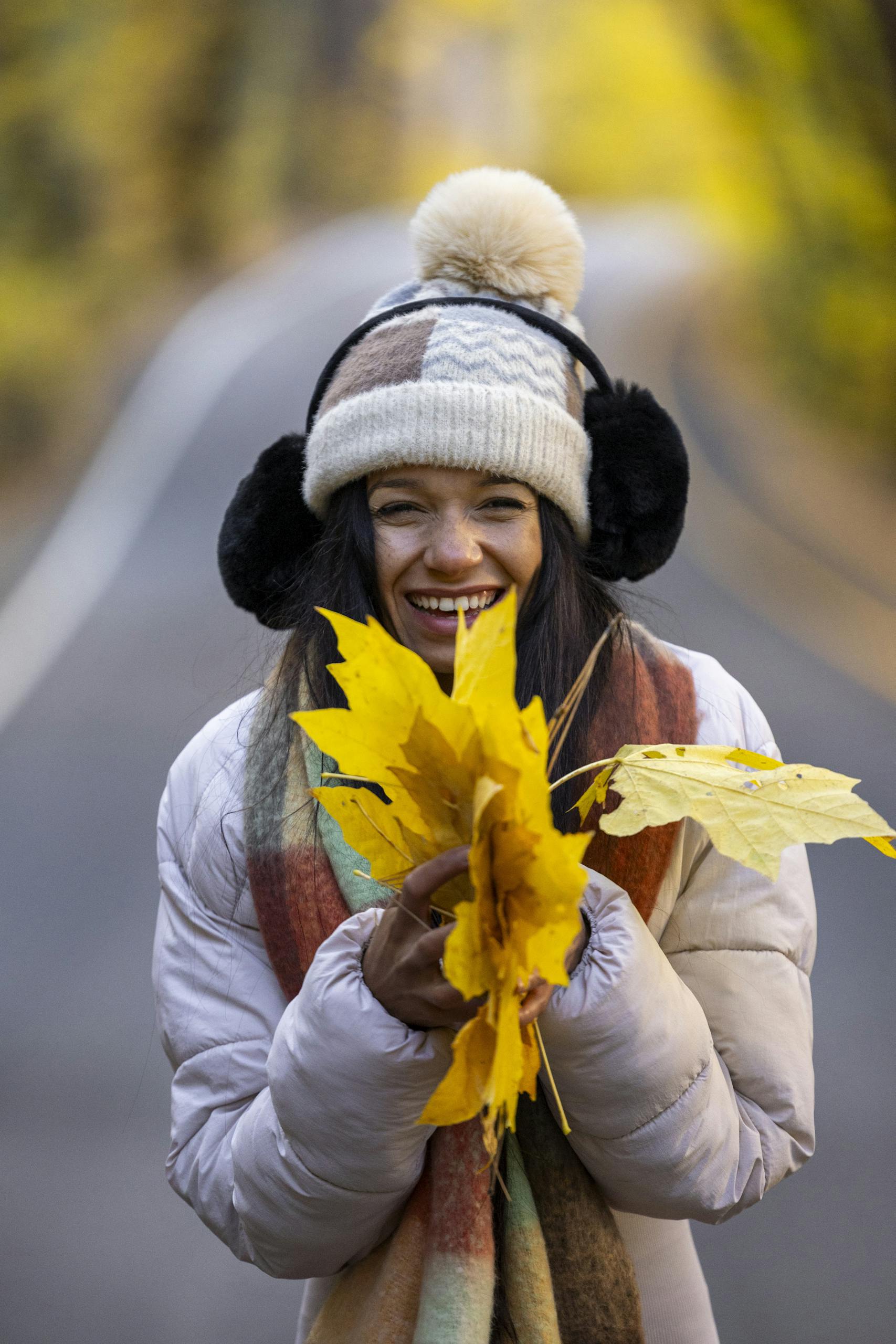 Smiling woman in cozy attire holding yellow leaves on a scenic fall day outdoors.