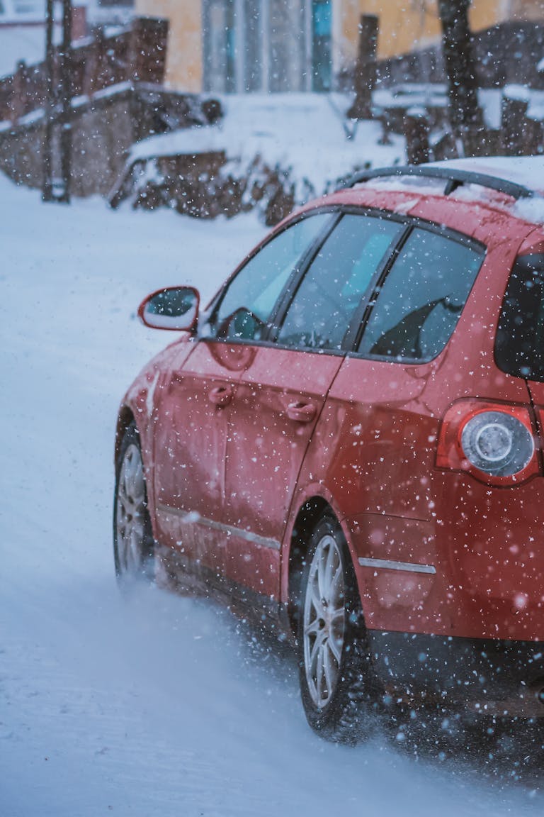 A red car navigates a snowy urban street during winter, showcasing a serene cityscape.