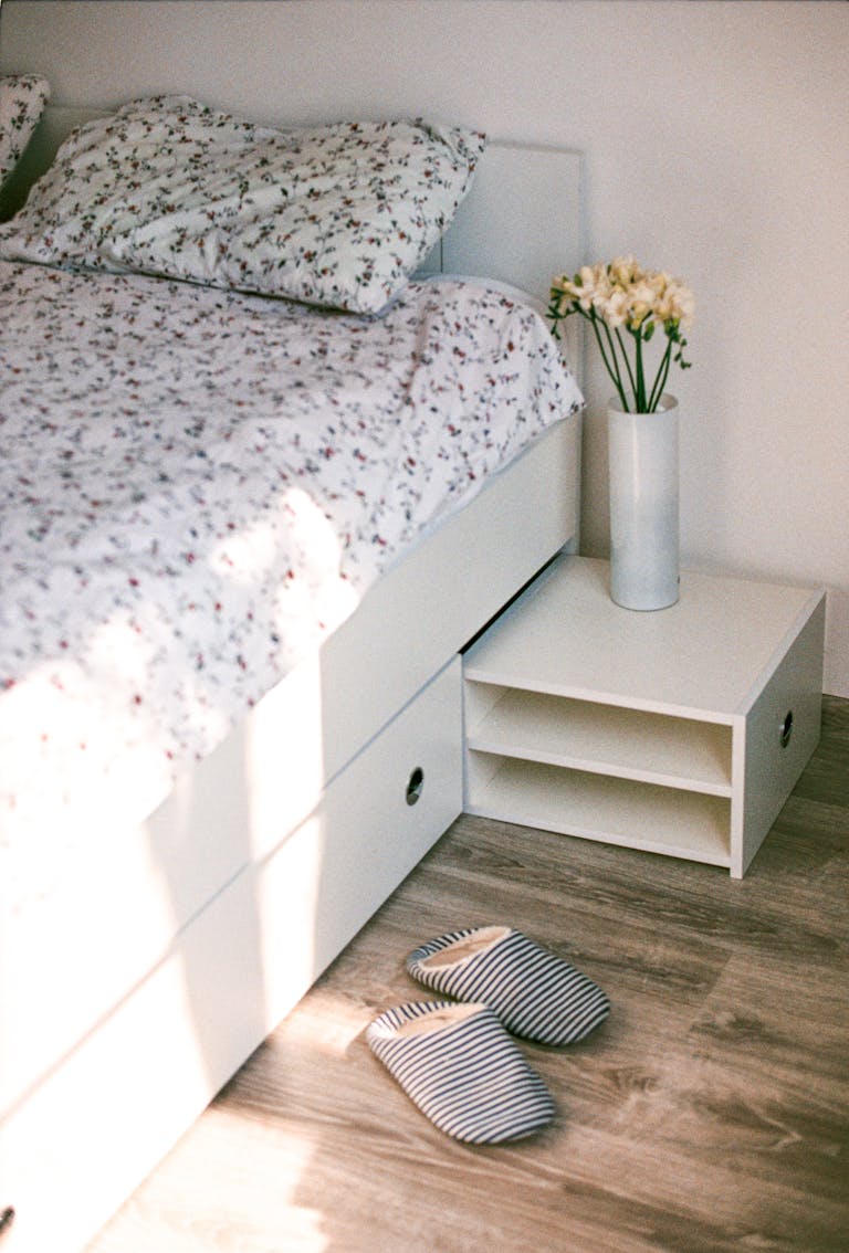 A warm and inviting bedroom featuring a floral-patterned bed, a nightstand with flowers, and striped slippers on wooden floor.