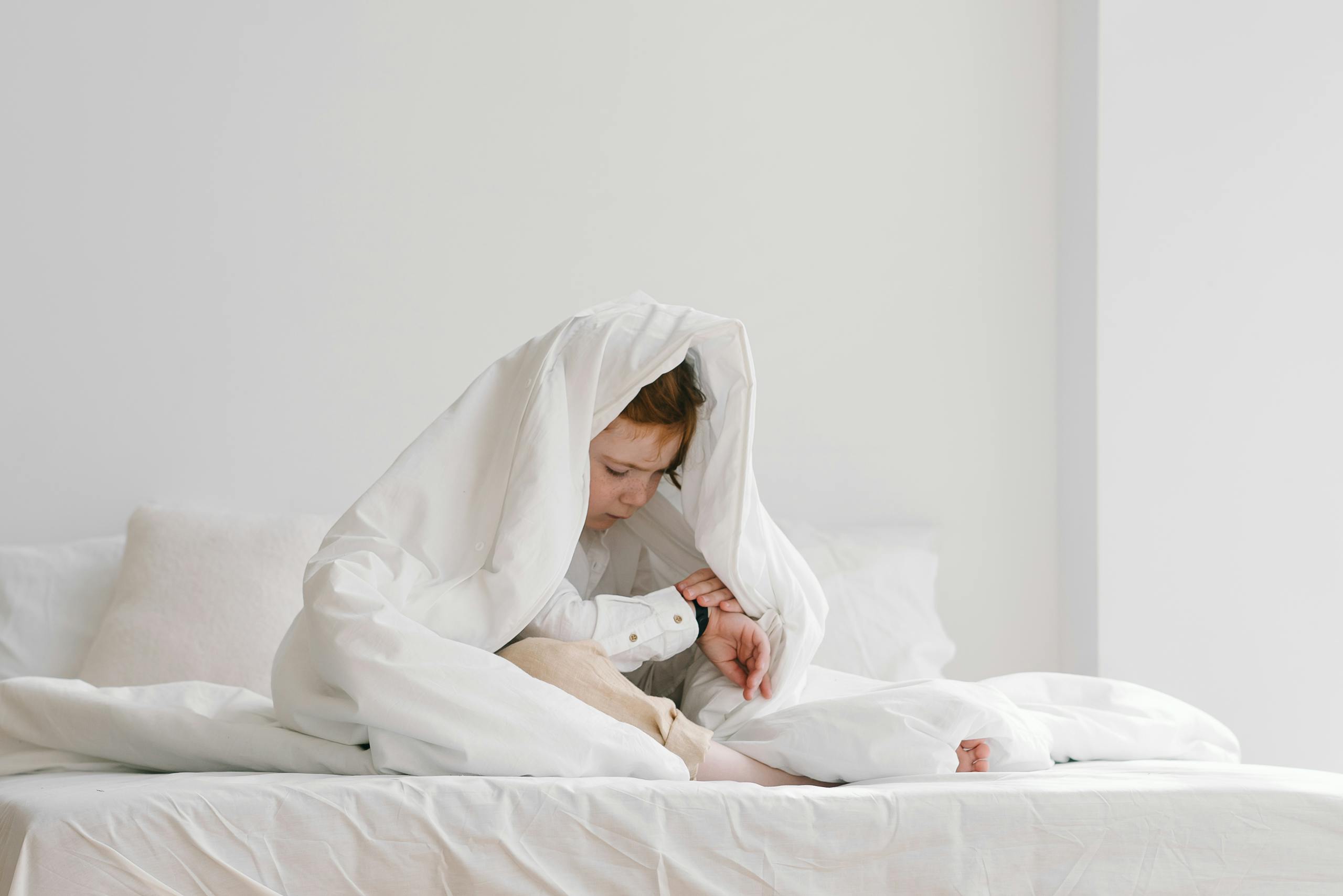 A young child sitting playfully under a white duvet on a bed in a bright room.
