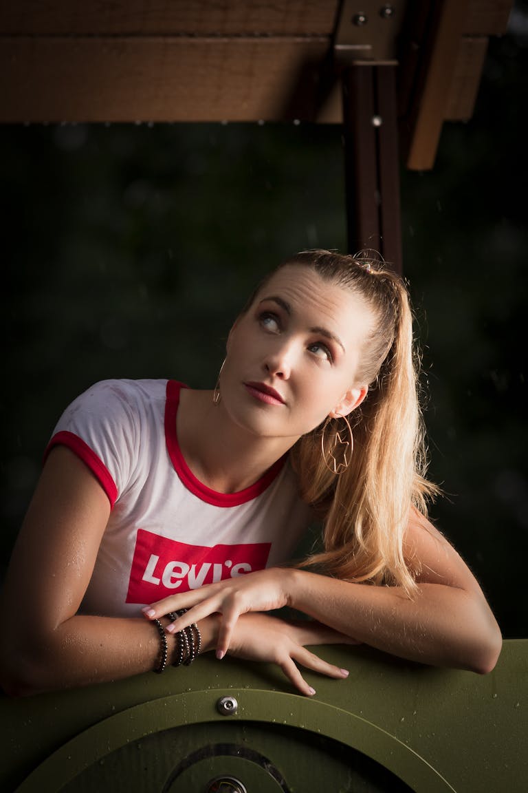 Young woman with blonde hair poses thoughtfully under a shelter looking upwards.