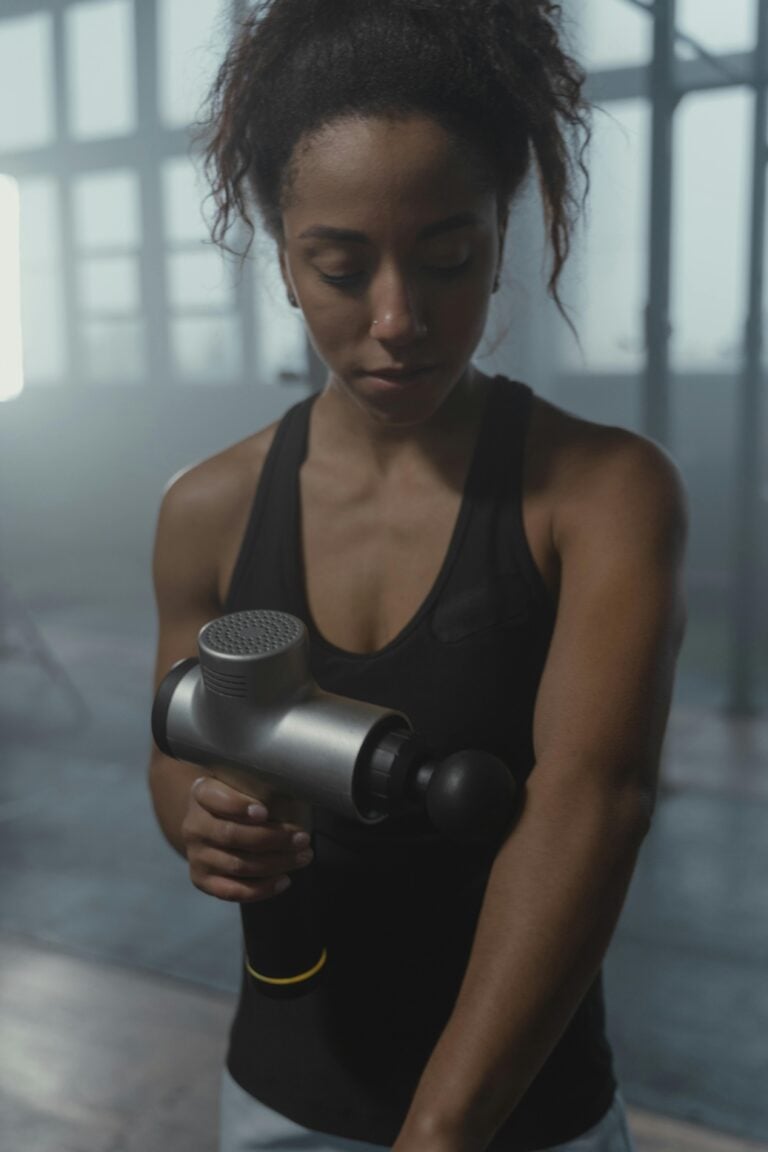 A focused woman using a massage gun on her arm in a gym environment.