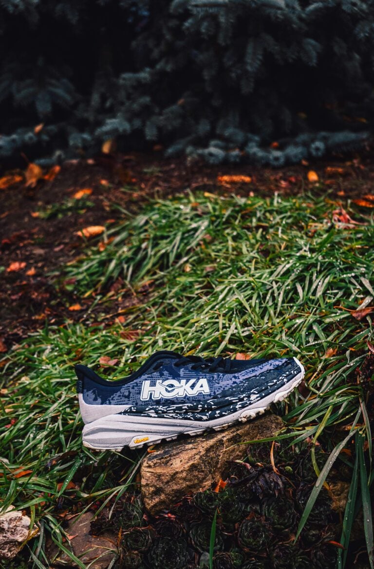 Outdoor trail running shoe resting on a rock surrounded by lush greenery.