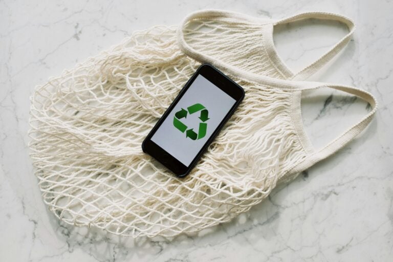 Top view of smartphone with recycling sign on screen placed on white textile mesh bag on white marble table in light room