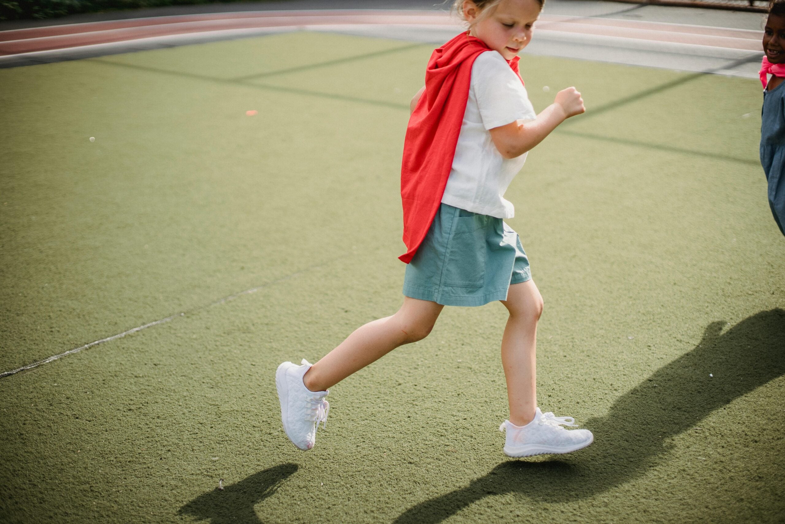A cheerful child wearing a red cape running on a sports field on a sunny day.