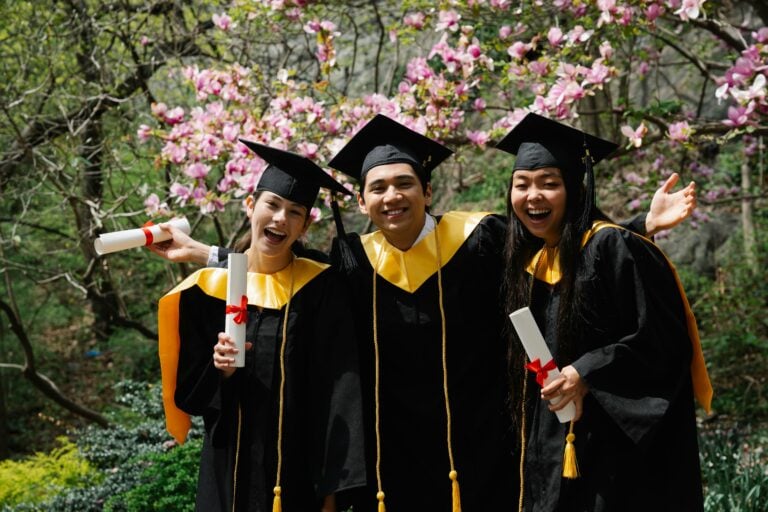 Three graduates celebrate outdoors with diplomas under flowering trees.