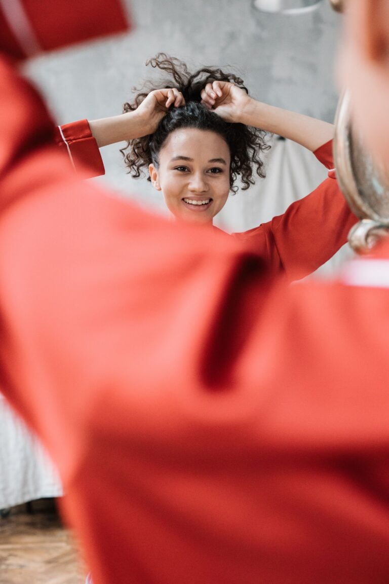 Woman in red sleeves smiling while fixing hair in front of mirror indoors.