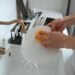 Hands washing a white dish in a modern kitchen sink with a yellow sponge.