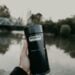 A hand holds a Stanley vacuum bottle near a serene riverside, perfect for outdoor and travel themes.
