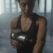 A focused woman using a massage gun on her arm in a gym environment.