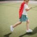 A cheerful child wearing a red cape running on a sports field on a sunny day.