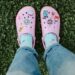 Bright pink crocs adorned with stickers, worn with blue socks, on lush green grass.